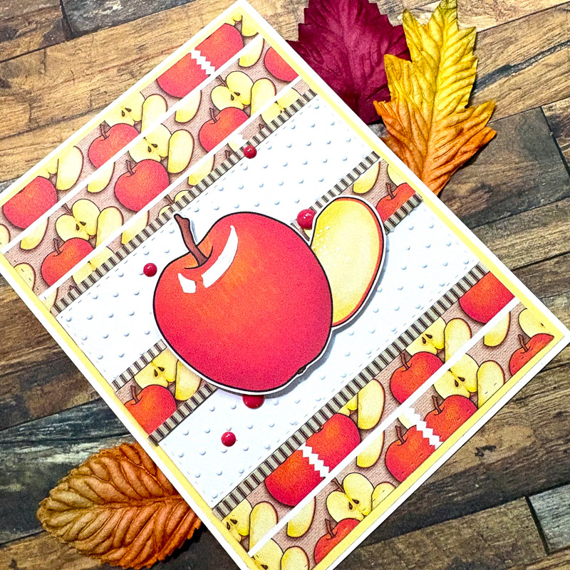 hand-made greetings card featuring a bright red apple in the centre and a smaller apple pattern surrounding, set of a wooden table with autumn leaves