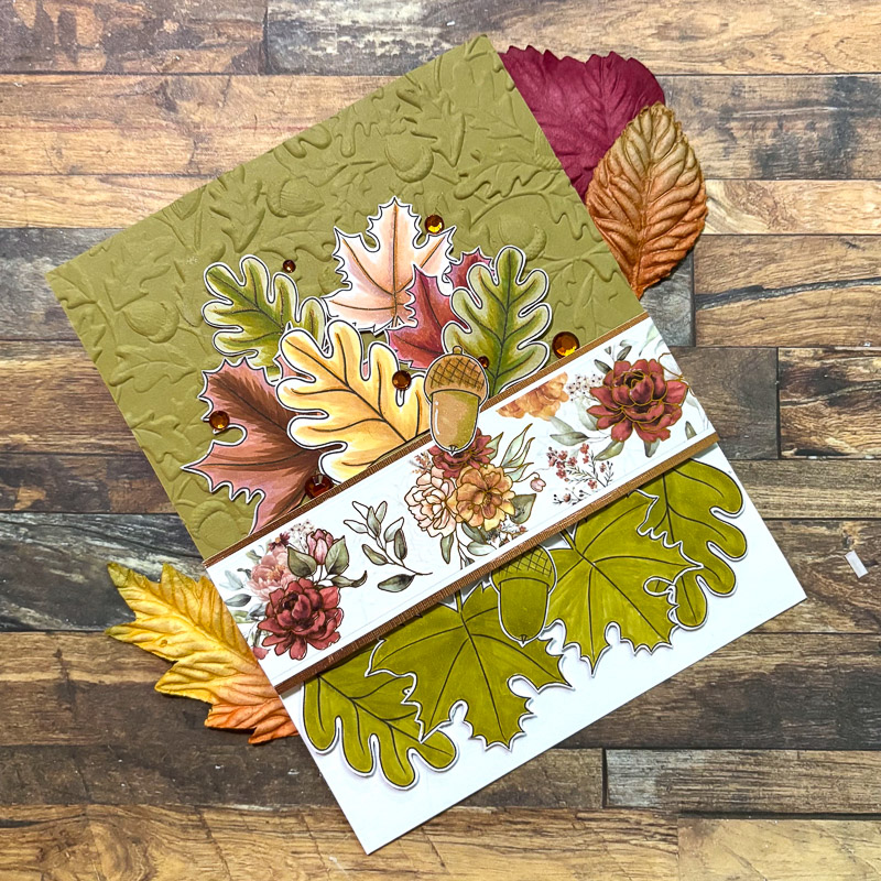 hand-made greetings card featuring a decorative border and hand coloured autumn oak leaves and acorns on a wooden table top
